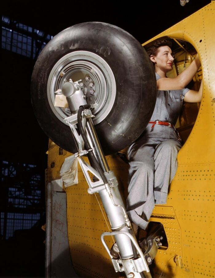 Woman at work during WWII in a factory wearing coveralls, inspecting an airplane's landing gear inside a hangar.