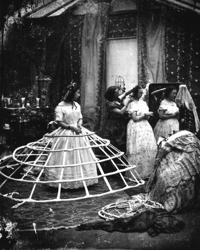Women in 1800s America preparing dresses with hoop skirts and styling hair in a vintage indoor setting.
