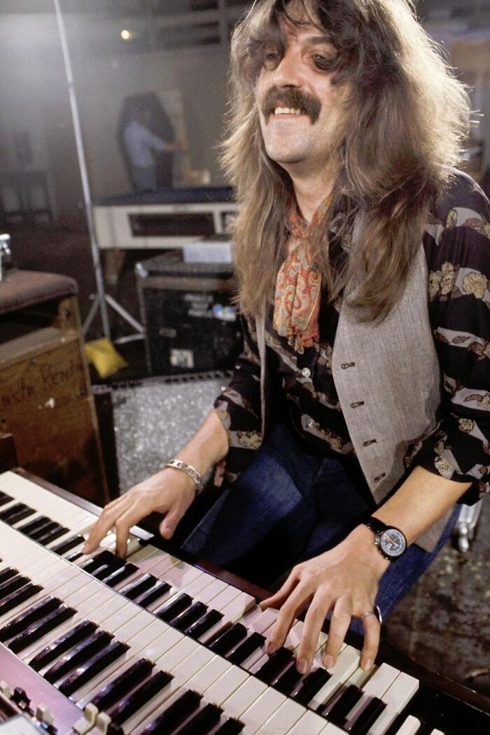 1970s rock legend playing keyboard in a studio, wearing vintage clothing and smiling during music creation session.