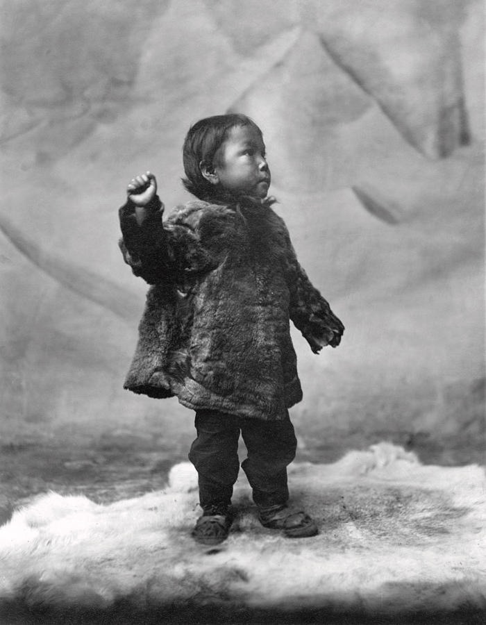 Inuit child wearing traditional fur clothing standing on fur, showcasing the unbreakable spirit in a historic black and white photo.