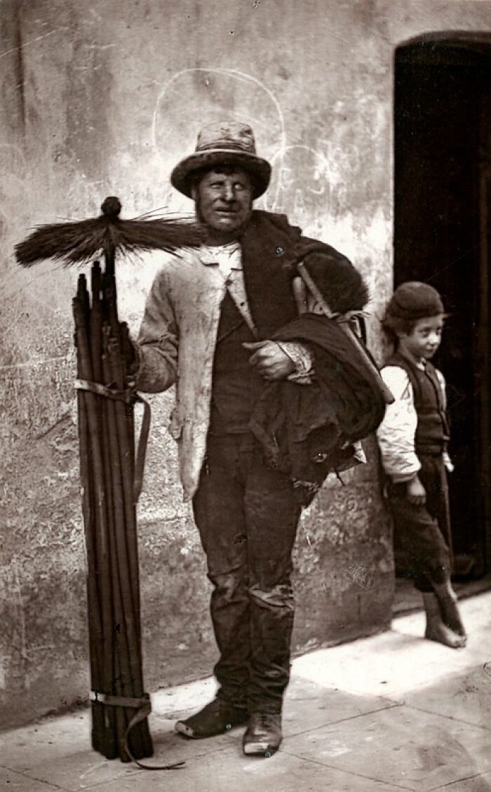Man in worn clothes holding brooms, standing against wall, illustrating how London's poorest lived in the 1800s.