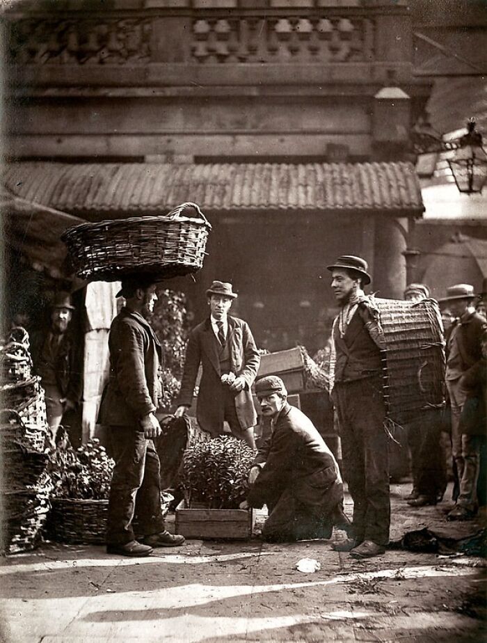 London's poorest workers in the 1800s carrying baskets and crates in a crowded urban outdoor market scene.