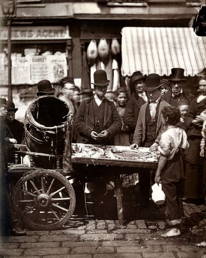 Street scene showing London’s poorest people in the 1800s gathered around a small market table near a wooden cart.