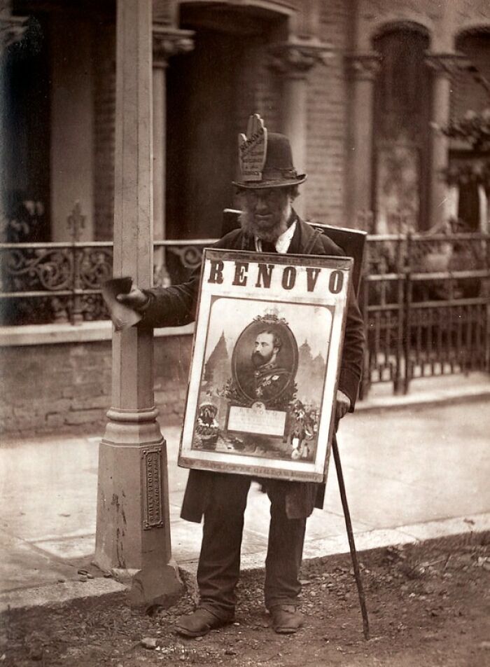 Victorian man representing London's poorest living in the 1800s, standing on a street with a sign and cane.