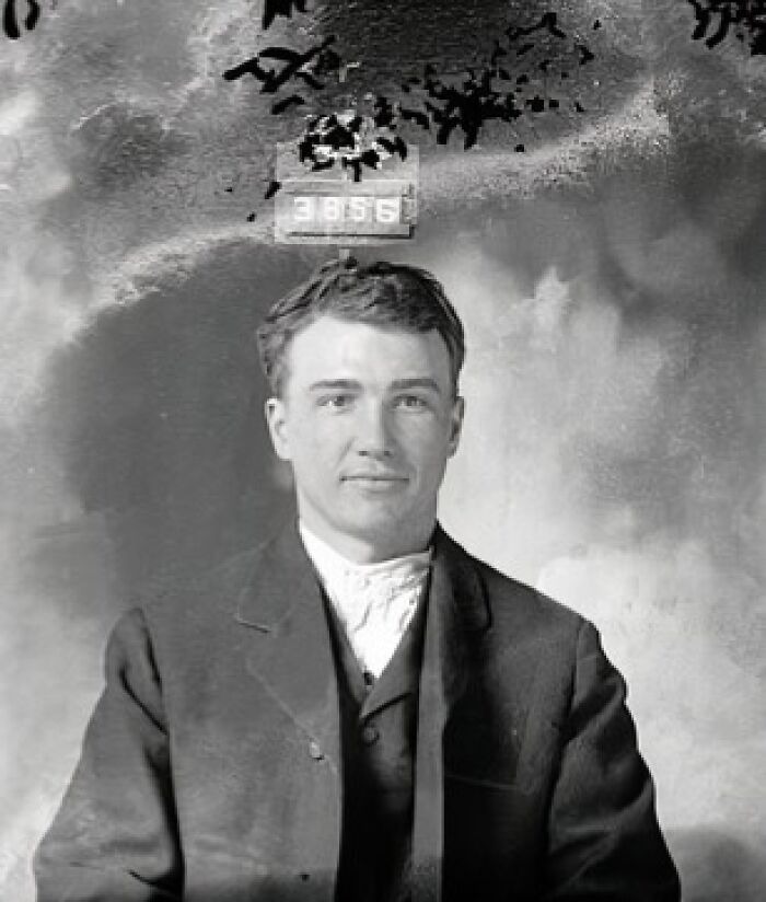 Young man in a historical mugshot wearing vintage suit and holding arrest identification number in black and white photo.