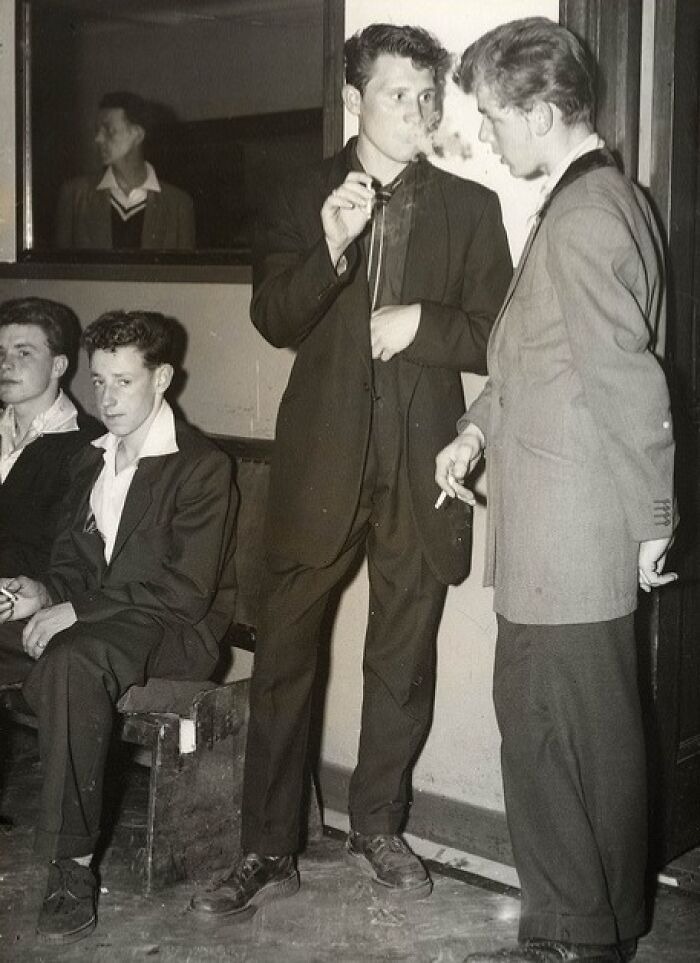 Black and white photo of 20th century Teddy Boys in suits smoking indoors, showcasing vintage fashion and youth culture.