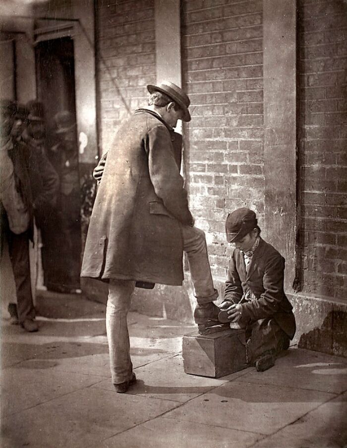 London's poorest in the 1800s with a boy shining shoes on a city street beside a brick wall.
