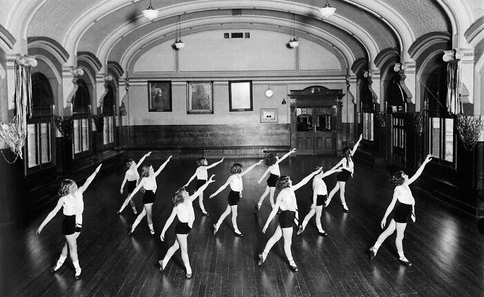 Black and white historical photograph of young girls practicing gymnastics in a large hall from 100 years ago.