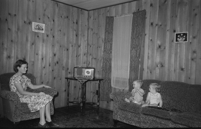 Woman in a floral dress sitting on a chair listening to a radio while two toddlers sit on a couch in a Great Depression era room