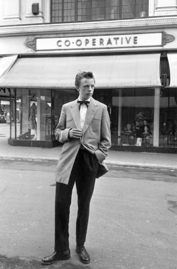 Young man in classic Teddy Boy era outfit standing on street in front of Co-operative store, 20th century fashion style.