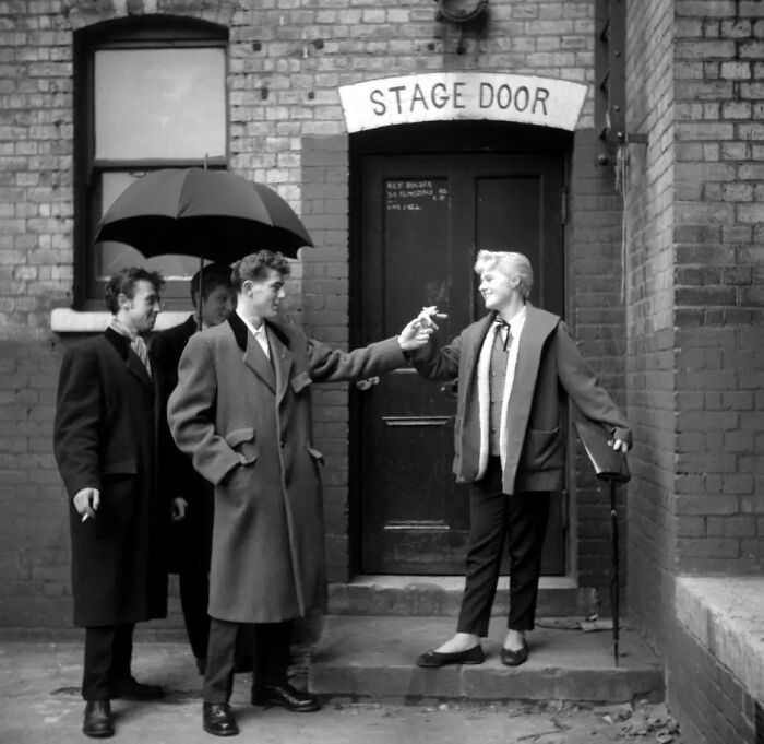 Group of 20th century Teddy Boys dressed in long coats and scarves, socializing outside a stage door on a city street.