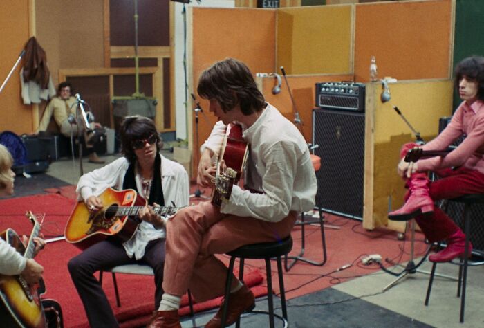 1970s rock legends playing acoustic guitars in a vibrant studio, capturing raw moments of music creation.