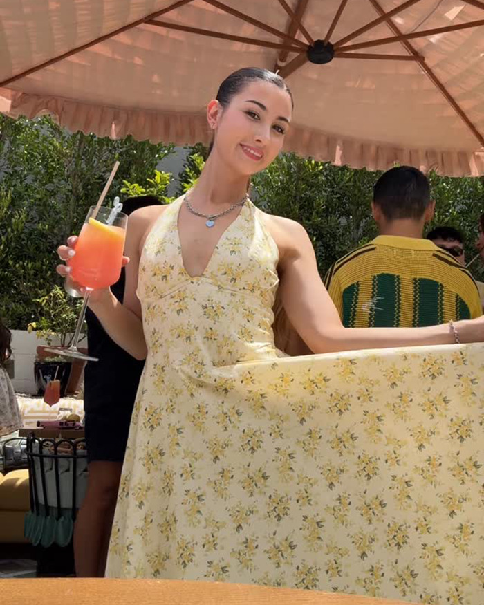 Young woman enjoying a drink outdoors in a floral dress under an umbrella, illustrating influencer travel hack backfiring.