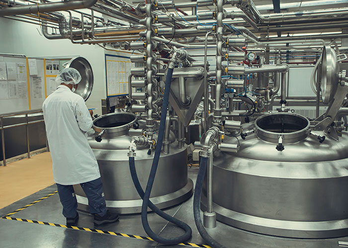 Worker in protective gear inspecting large industrial tanks and pipes in a sterile factory setting for safety compliance.