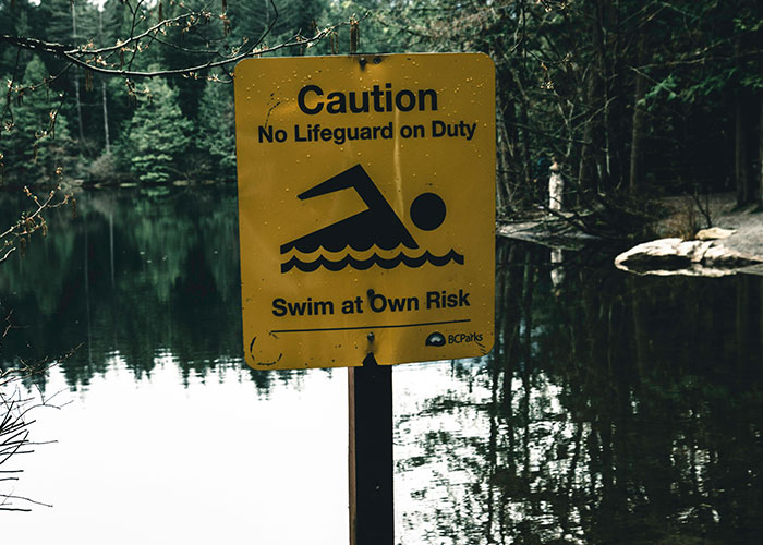 Yellow caution sign about no lifeguard and swimming at own risk near a dark, eerie lake in a forest setting, creepy true stories.