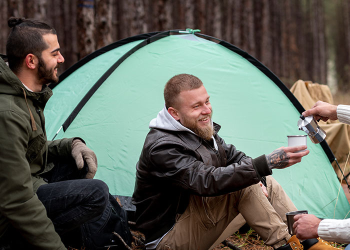Two men enjoying coffee by a green tent in the forest, capturing casual moments in true creepy stories setting.