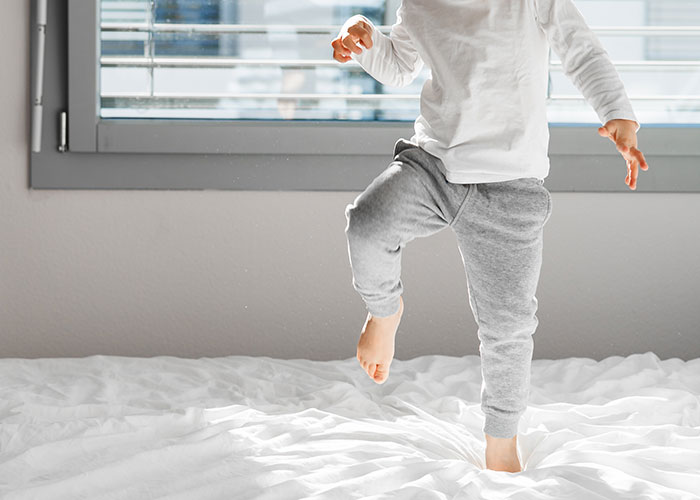 Child in gray pants and white shirt stepping on a bed in a bright room, evoking creepy true stories atmosphere.
