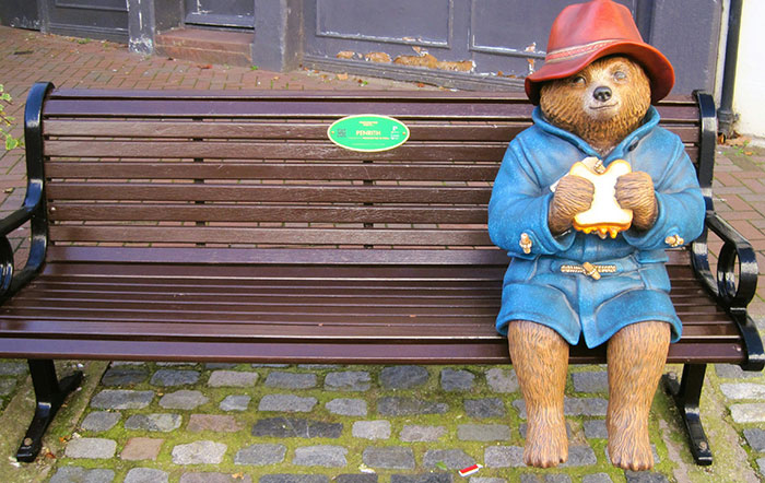 Paddington Bear statue sitting on a bench wearing a blue coat and red hat, a cool fact about the world in June.