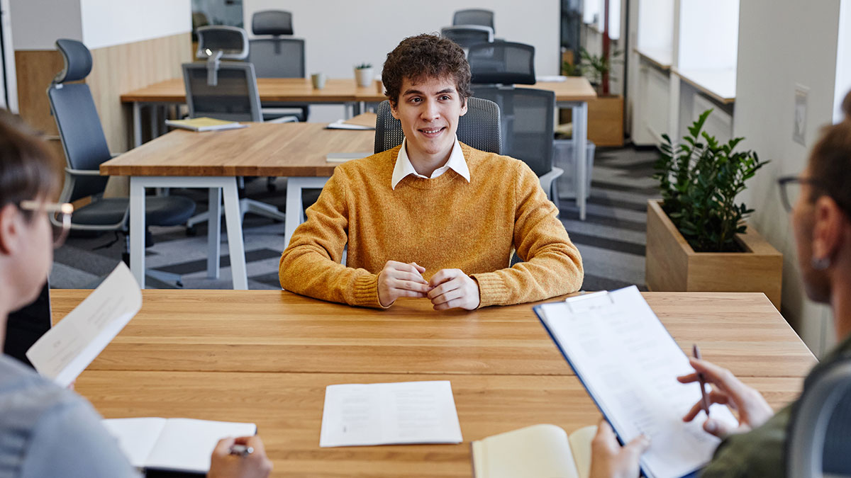 Young man in a job interview sitting at a table with two interviewers reviewing documents in a modern office setting