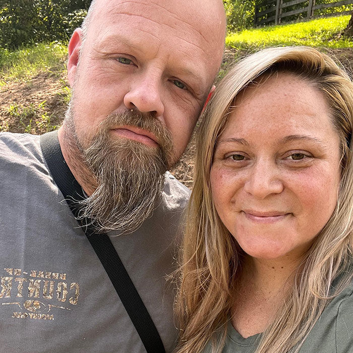 Middle-aged man with beard and woman with blonde hair smiling outdoors near a sunny green area in casual clothes.