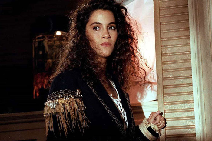 Jami Gertz with voluminous curly hair wearing a black jacket with fringe and bead details in an indoor setting.