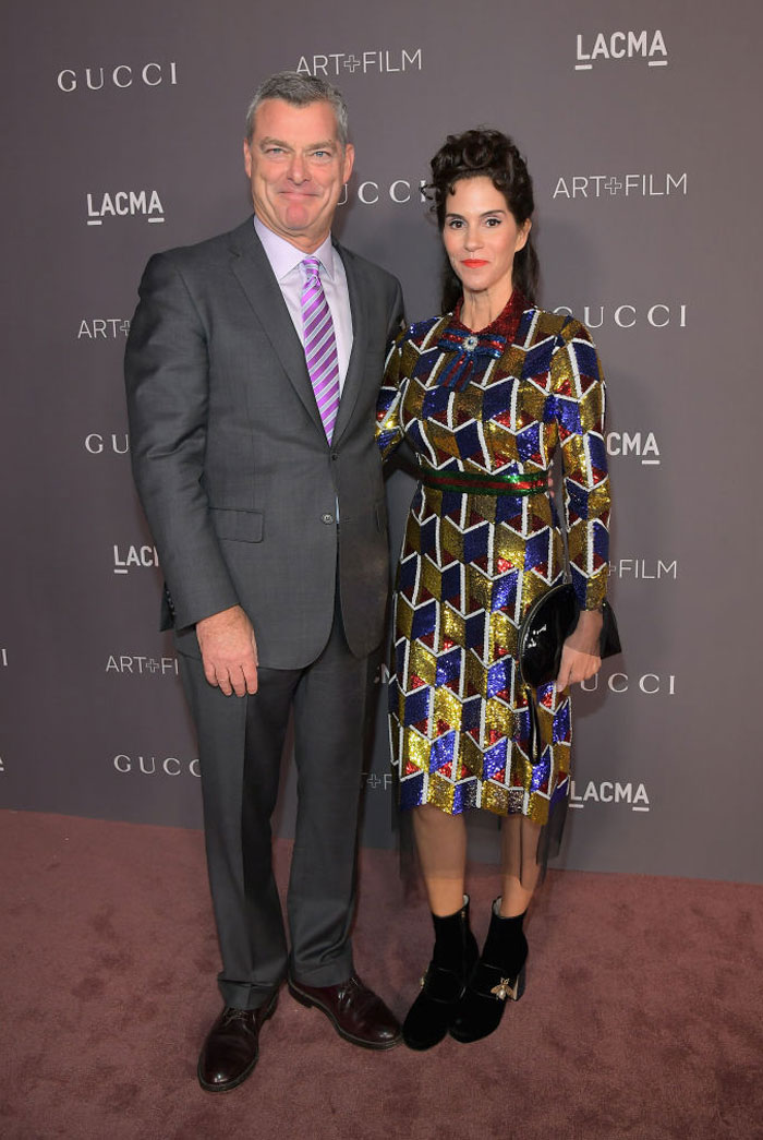 Jami Gertz at a formal event wearing a colorful geometric dress, standing next to a man in a gray suit and purple tie.
