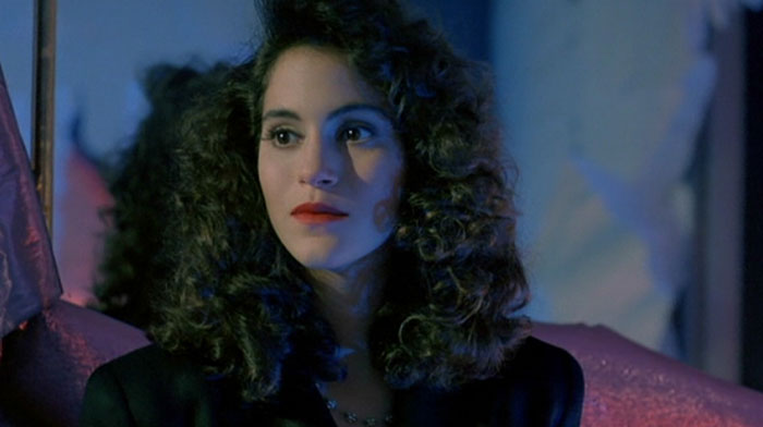 Jami Gertz with curly hair and red lipstick, wearing a dark outfit in a dimly lit indoor setting.