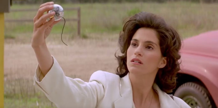 Jami Gertz outdoors holding a small object, wearing a white jacket with a thoughtful expression, under the radar success.