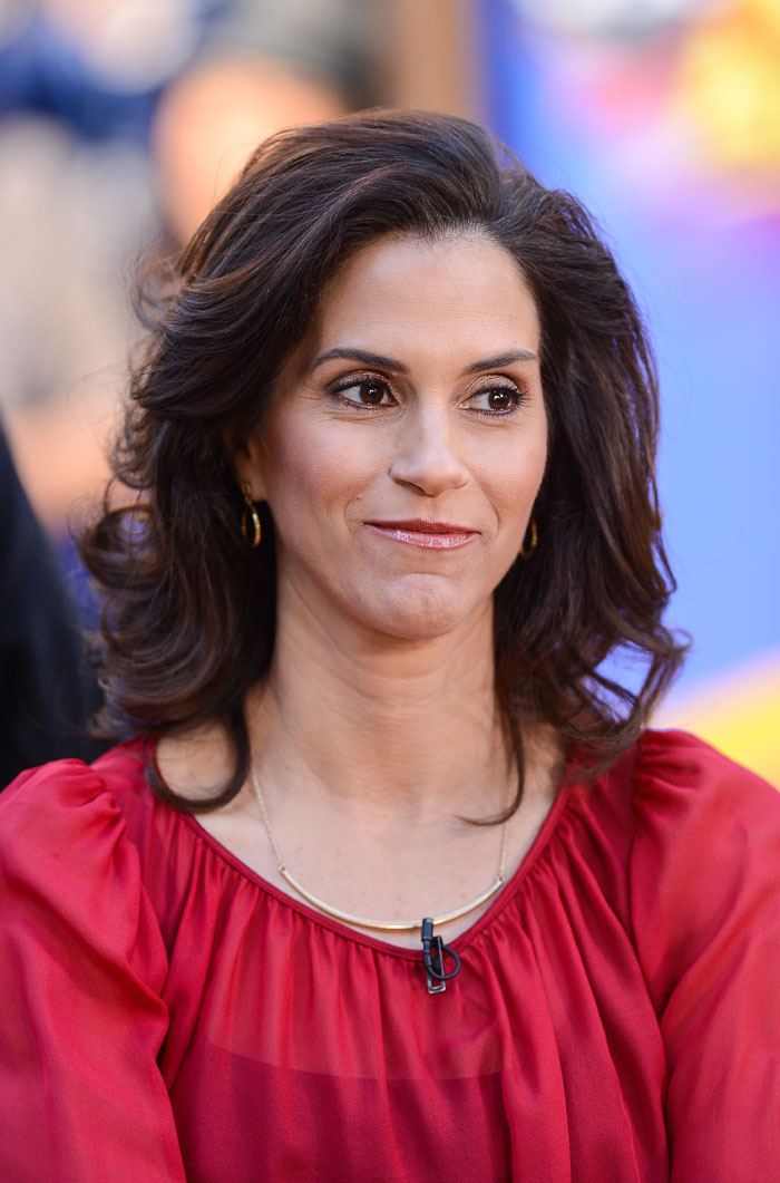Jami Gertz wearing a red blouse and gold earrings, smiling slightly with styled brown hair in a blurred background.