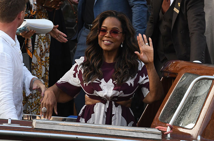 Oprah with long hair and sunglasses waving while standing on a boat, representing Jami Gertz success under the radar.