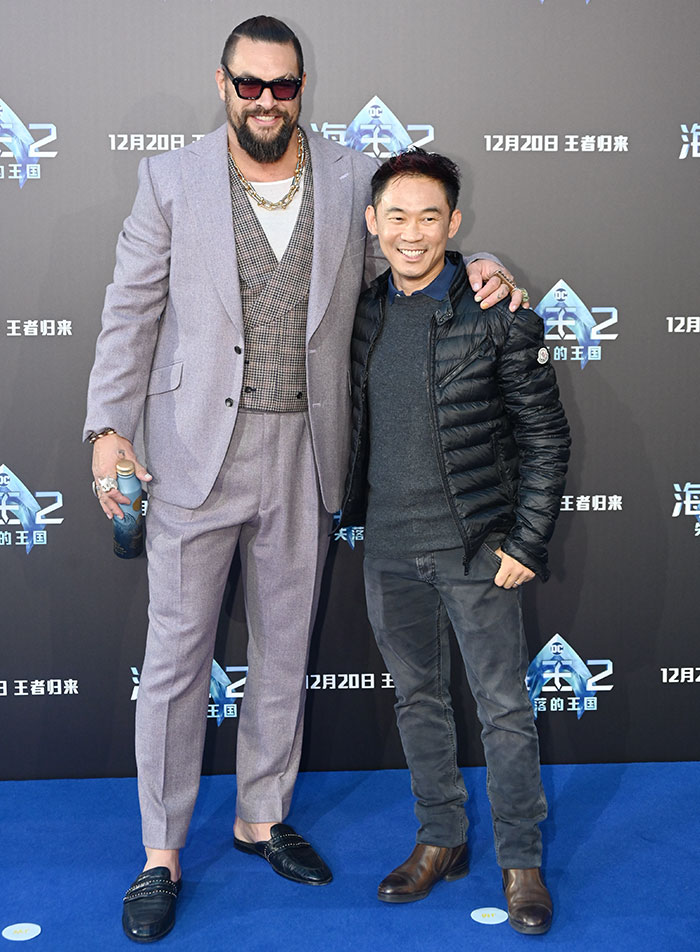 Tall celebrity wearing a lavender suit and sunglasses standing next to a shorter man in casual black jacket at a movie premiere.