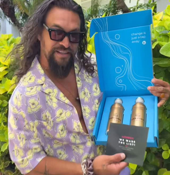 Jason Momoa without iconic beard holding a blue box with two gold bottles outdoors near greenery. - 2