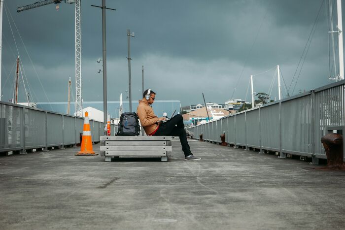 Person who ran away from home sitting alone on a bench at a pier, wearing headphones and using a laptop on a cloudy day. - 31