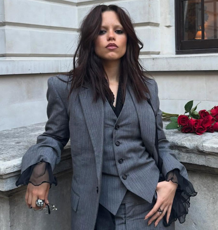 Young woman in a gray pinstripe suit with sheer sleeve accents poses confidently by stone ledge with red roses nearby Young woman in a gray pinstripe suit with sheer sleeve accents poses confidently by stone ledge with red roses nearby