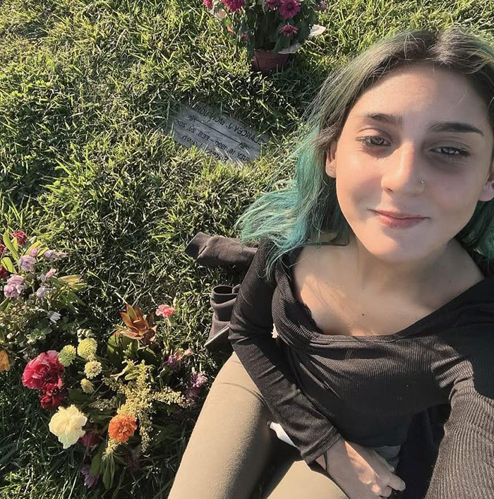 Young woman with teal hair sitting by a grave with flowers, related to 7-Eleven worker passing away after manager incident.
