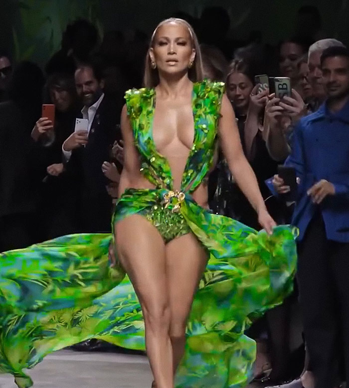 Jennifer Lopez walking runway in a green flowing dress, facing forward during a raunchy show with crowd capturing the moment.