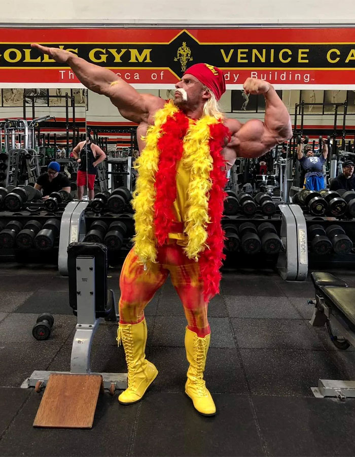Joey Swoll posing in a gym with muscles flexed, dressed in a Hulk Hogan-inspired outfit and feathered scarf.