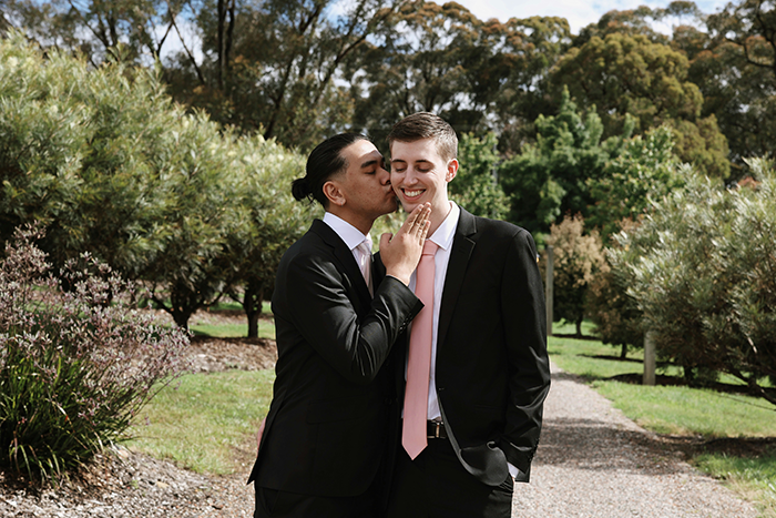 Gay couple dressed in suits sharing a tender moment outdoors in a park with trees and greenery around.