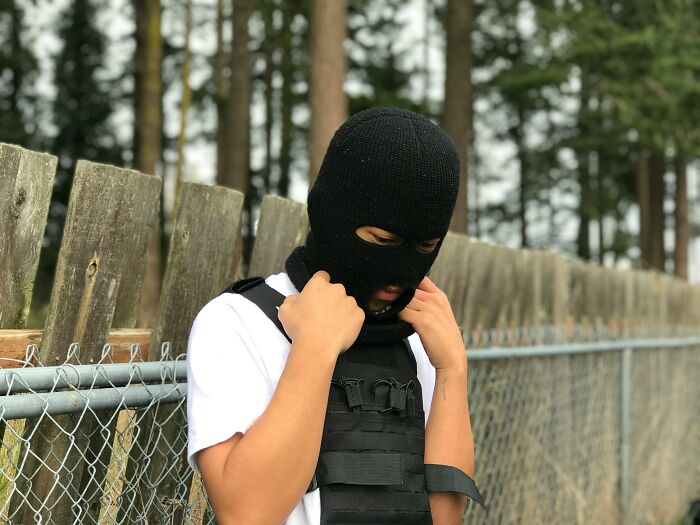 Person wearing a black ski mask and tactical vest standing outside near a wooden fence, reflecting bizarre McDonald's employee experiences.