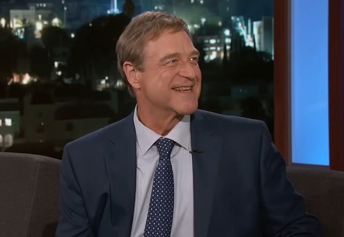 John Goodman on a talk show set, wearing a suit and tie, smiling and showcasing his weight loss transformation.
