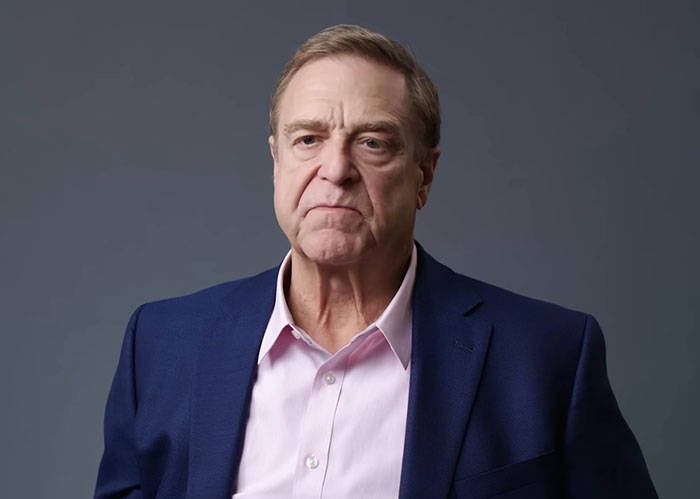 John Goodman in a navy blazer and light pink shirt, showing off his drastic 200-lb weight loss against a gray background.