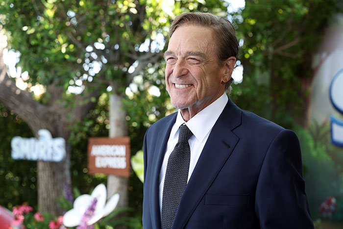 John Goodman smiling outdoors in a suit, showcasing his drastic 200-lb weight loss transformation.