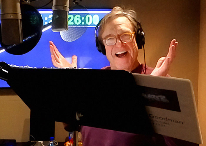 John Goodman in a recording studio, wearing headphones and glasses, showing off his drastic 200-lb weight loss.