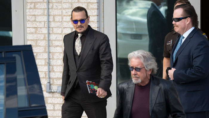 Johnny Depp wearing a black suit and sunglasses, walking outside with two men in dark suits near a building.