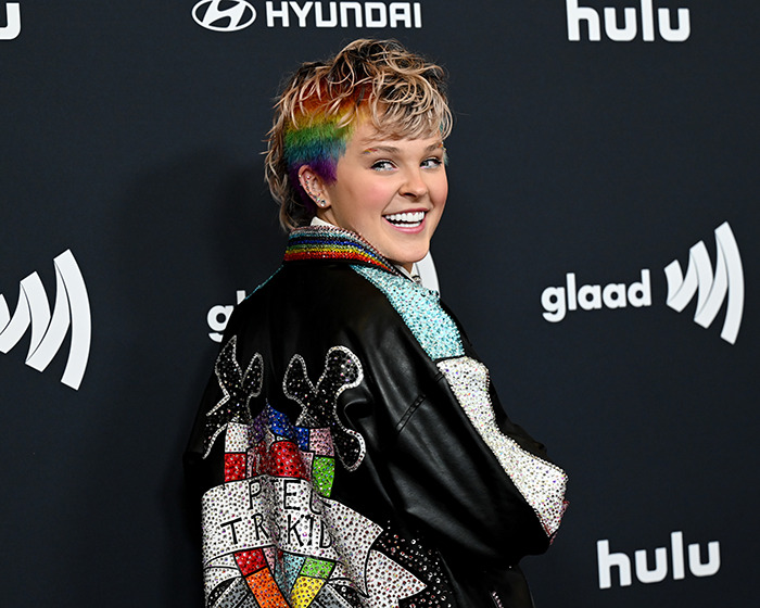 Person with short, rainbow-colored hair wearing a studded jacket, representing JoJo Siwa's straight pop trad wife transformation.