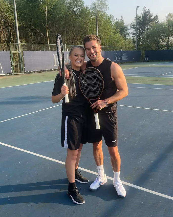 Two people smiling on a tennis court holding rackets, capturing a moment related to straight pop genre and JoJo Siwa.