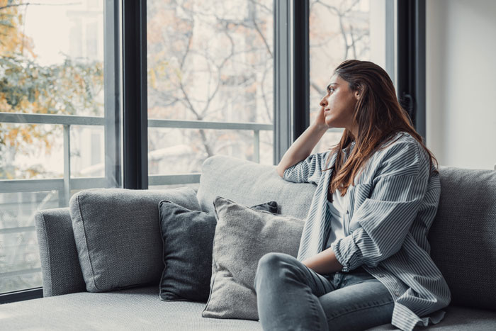 Woman sitting on couch looking out window, reflecting after boyfriend’s family made a scar joke at gathering. Woman sitting on couch looking out window, reflecting after boyfriend’s family made a scar joke at gathering.