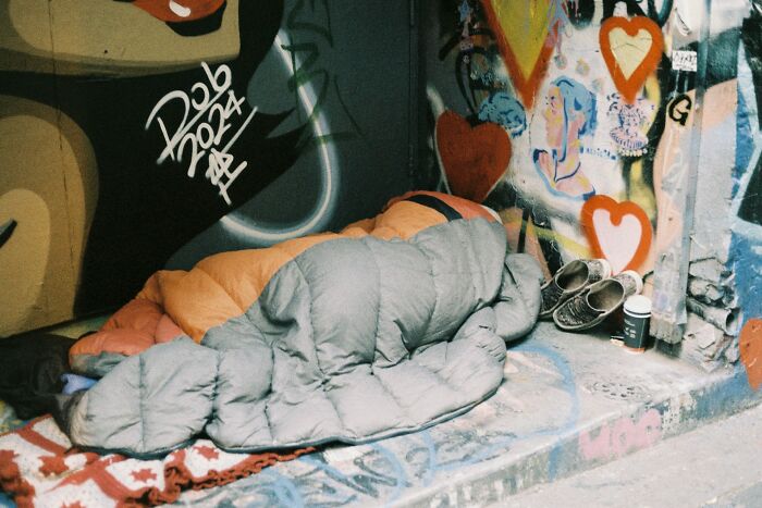 Person sleeping on a sidewalk wrapped in a sleeping bag near graffiti, illustrating harsh punishment by parents leaving kids alone.