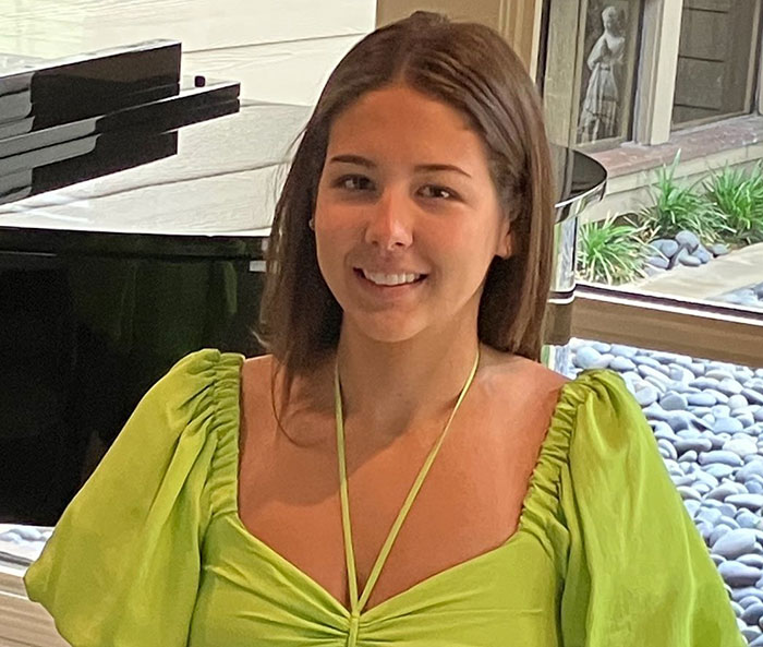 Young flood victim smiling indoors near a piano and window with rocks and greenery outside.