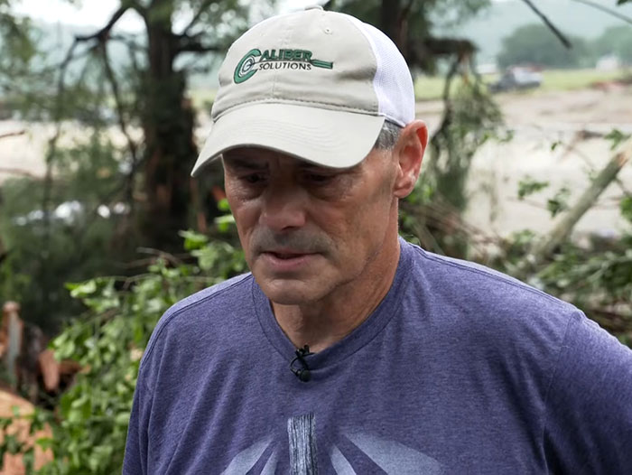 Man wearing a Capiber Solutions hat looks concerned, standing outdoors near flood-damaged area reflecting flood victim fear.